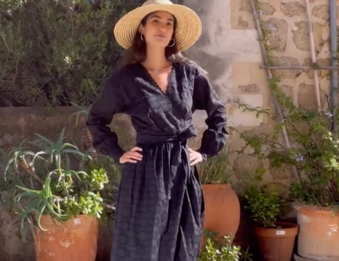 A woman wearing a long black dress with long sleeves, styled with a wide-brimmed hat and heels, standing in front of a rustic background with potted plants.