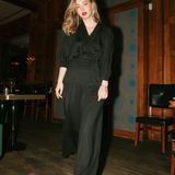 Woman in a black outfit standing in a dimly lit room with wooden walls and furniture.