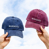 Two baseball caps held up by hands against a cloudy sky: one navy blue cap with the word 'MAMACITA' in white embroidery and one dusty burgundy cap with the word 'MOMAGER' in white embroidery.