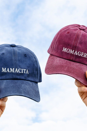 Two baseball caps held up by hands against a cloudy sky: one navy blue cap with the word 'MAMACITA' in white embroidery and one dusty burgundy cap with the word 'MOMAGER' in white embroidery.