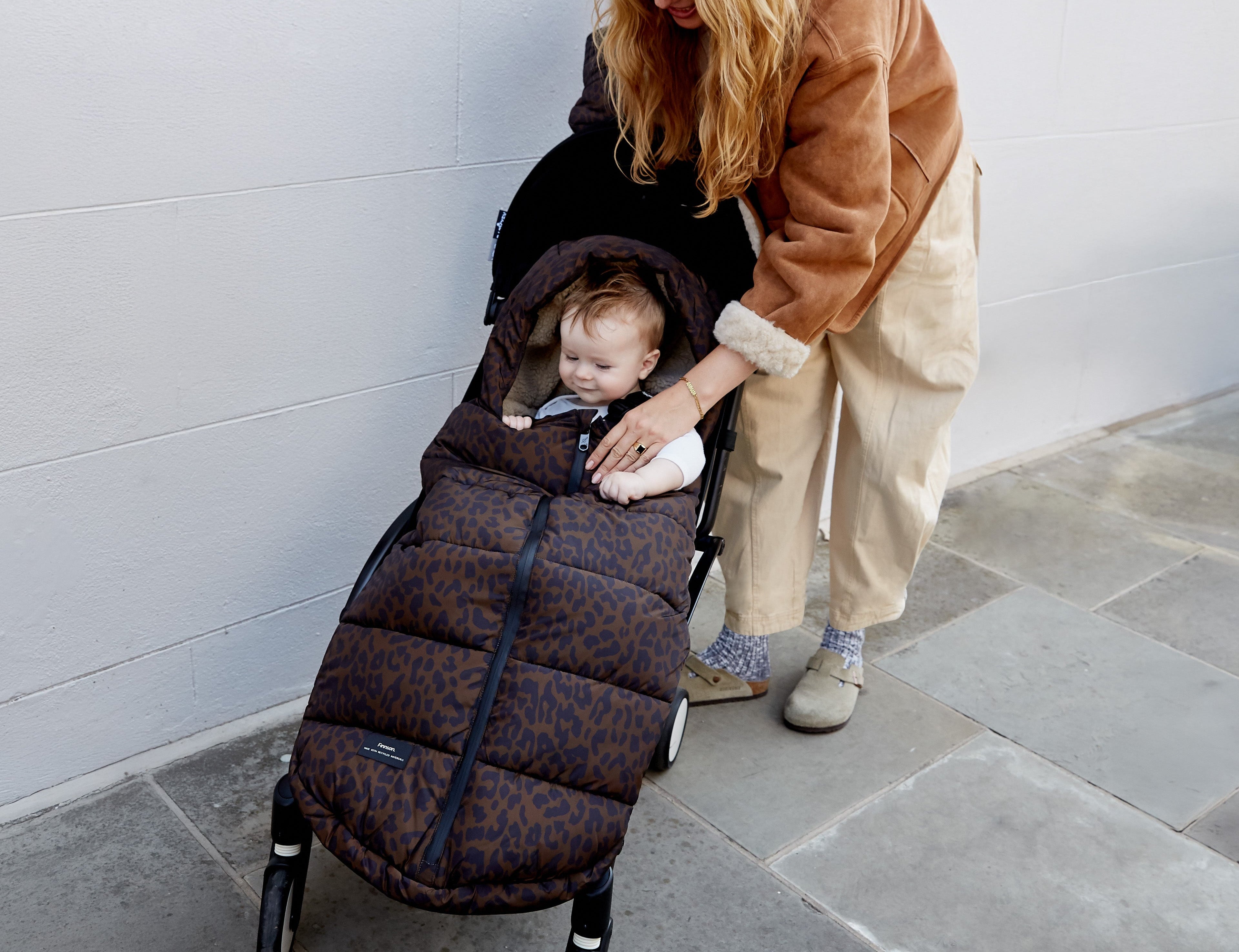A woman in a brown jacket and beige pants pushes a stroller while a baby sits inside wearing a dark brown patterned quilted baby liner. The scene is set outdoors against a light gray tiled wall and stone floor.