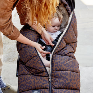 A baby stroller covered with a brown and black leopard print quilted winter blanket, with a woman adjusting the zipper while a baby sits inside.