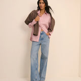 Woman wearing a pink striped shirt, brown jacket, and blue jeans against a beige background