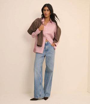 Woman wearing a pink striped shirt, brown jacket, and blue jeans against a beige background