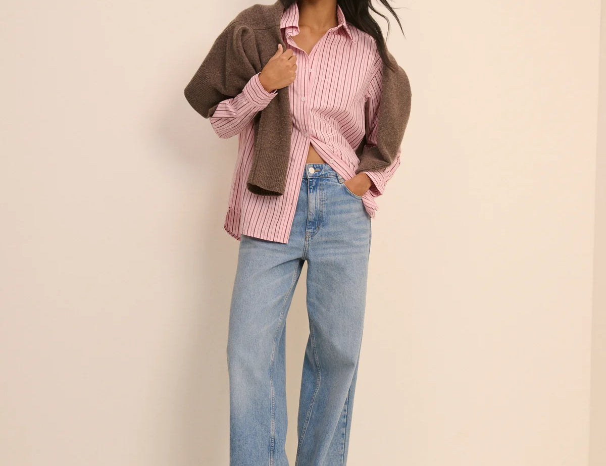 Woman wearing a pink striped shirt, brown jacket, and blue jeans against a beige background