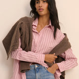 Woman wearing a pink striped shirt and blue jeans with a brown sweater over her shoulders against a beige background