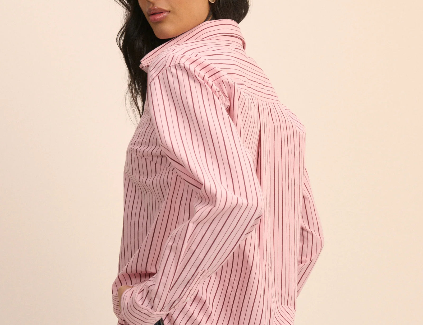 A woman wearing a pink and white vertically striped button-up shirt with a high collar, paired with light blue jeans, photographed against a solid light beige background.