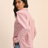 A woman wearing a pink and white vertically striped button-up shirt with a high collar, paired with light blue jeans, photographed against a solid light beige background.