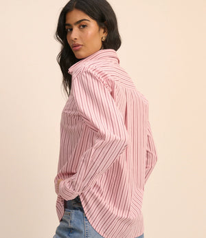 A woman wearing a pink and white vertically striped button-up shirt with a high collar, paired with light blue jeans, photographed against a solid light beige background.