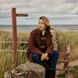 A woman sits on a large rock outdoors by the sea, wearing a brown coat, plaid shirt, blue jeans, and dark brown boots. A wooden signpost and grassy dunes are visible in the background under a cloudy sky.