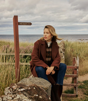A woman sits on a large rock outdoors by the sea, wearing a brown coat, plaid shirt, blue jeans, and dark brown boots. A wooden signpost and grassy dunes are visible in the background under a cloudy sky.