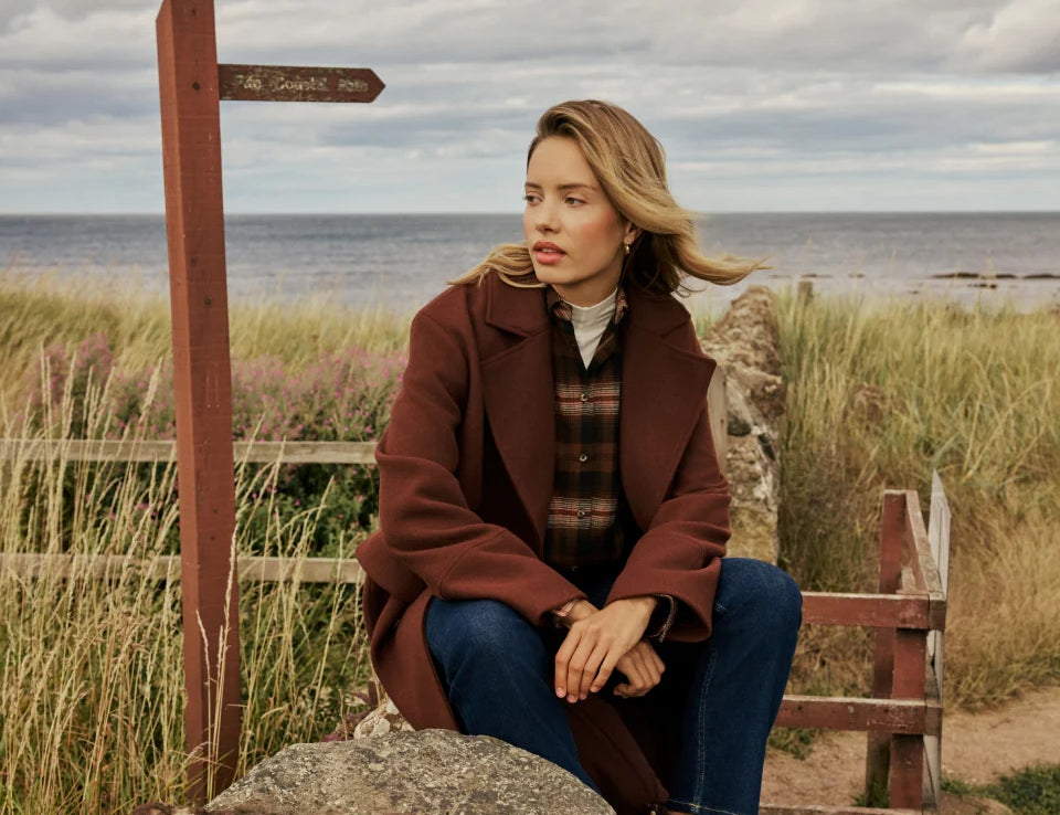 A woman sits on a large rock outdoors by the sea, wearing a brown coat, plaid shirt, blue jeans, and dark brown boots. A wooden signpost and grassy dunes are visible in the background under a cloudy sky.
