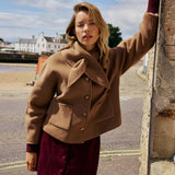 A woman wearing a camel brown cropped jacket and a burgundy corduroy skirt, leaning against a stone wall outdoors with a coastal town in the background.