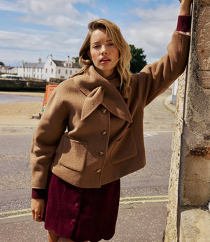 A woman wearing a camel brown cropped jacket and a burgundy corduroy skirt, leaning against a stone wall outdoors with a coastal town in the background.