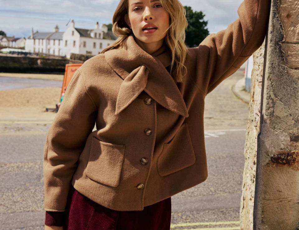 A woman wearing a camel brown cropped jacket and a burgundy corduroy skirt, leaning against a stone wall outdoors with a coastal town in the background.