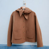 A camel brown wool coat with a large folded collar and decorative buttons, hanging on a wooden hanger against a light blue wall.
