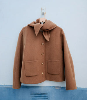 A camel brown wool coat with a large folded collar and decorative buttons, hanging on a wooden hanger against a light blue wall.