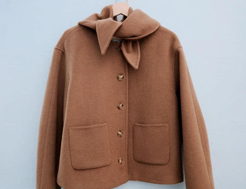 A camel brown wool coat with a large folded collar and decorative buttons, hanging on a wooden hanger against a light blue wall.