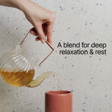 Tea being poured from a glass teapot into a pink cup with text 'A blend for deep relaxation & rest' on a speckled surface.
