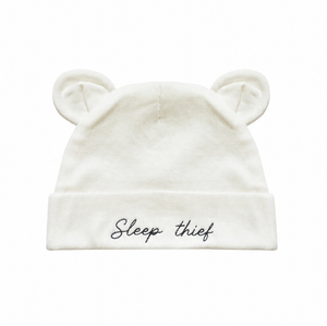 An off-white baby beanie hat with embroidered bear ears on top and the phrase 'Sleep thief' embroidered in black script on the front cuff, shown against a plain white background.