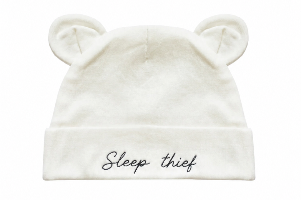 An off-white baby beanie hat with embroidered bear ears on top and the phrase 'Sleep thief' embroidered in black script on the front cuff, shown against a plain white background.