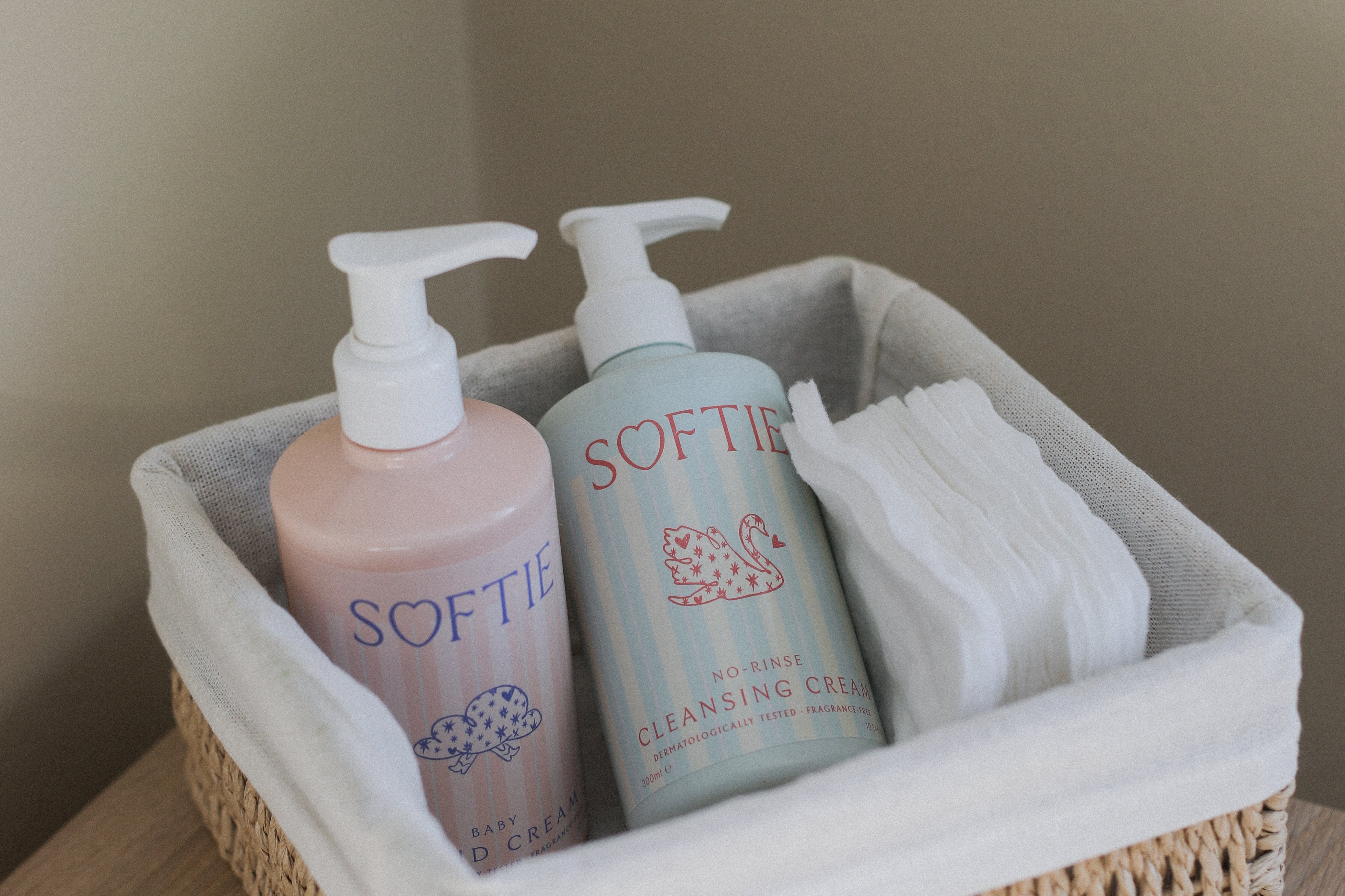 A woven natural fiber storage basket with a white fabric liner, holding two Softie pump bottles and a folded white towel, placed on a light wood surface against a beige wall.