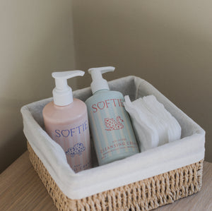 A woven natural fiber storage basket with a white fabric liner, holding two Softie pump bottles and a folded white towel, placed on a light wood surface against a beige wall.