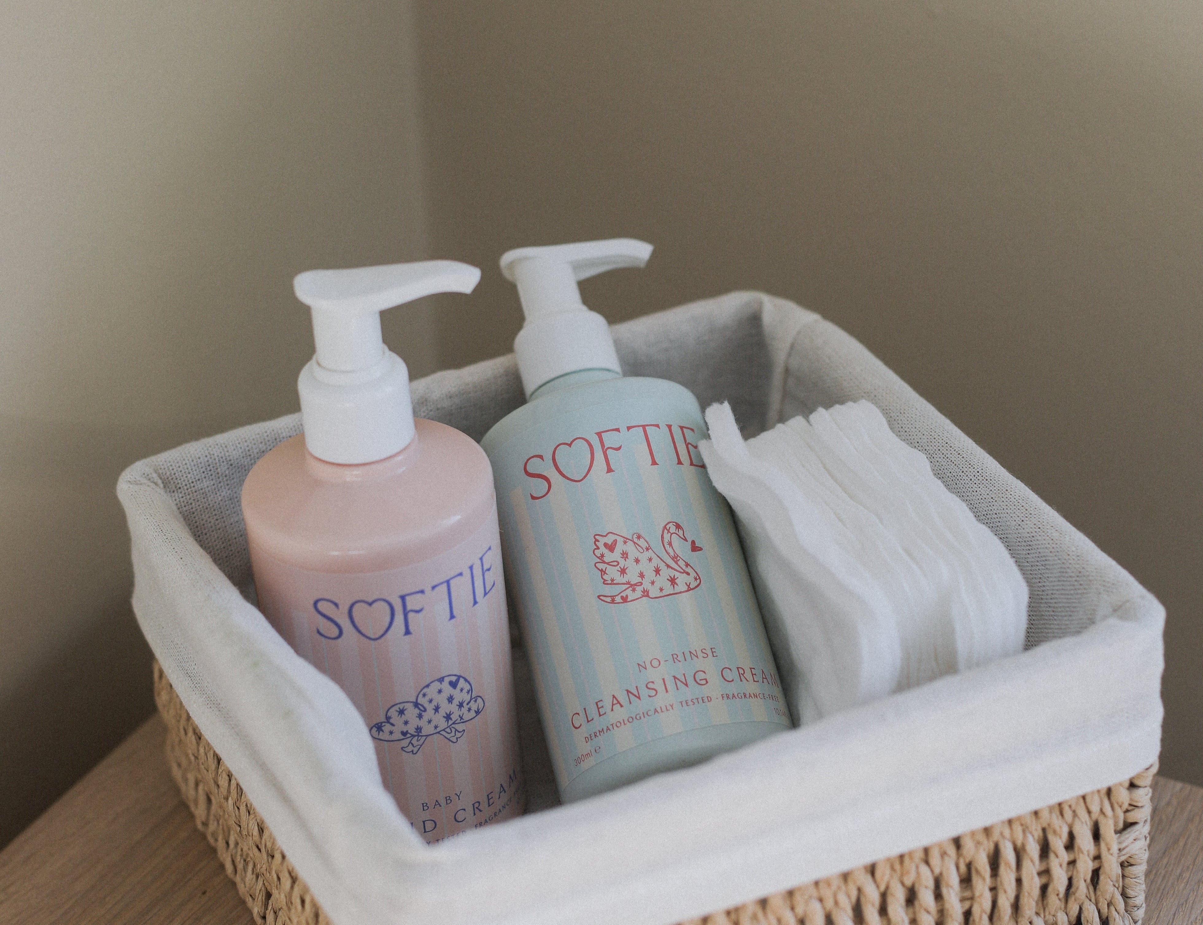 A woven natural fiber storage basket with a white fabric liner, holding two Softie pump bottles and a folded white towel, placed on a light wood surface against a beige wall.