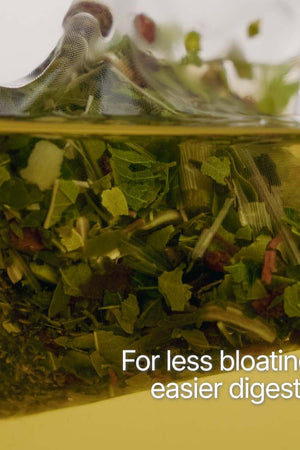 A clear tea bag filled with green tea leaves and herbs, partially submerged in yellow liquid with white text reading 'For less bloating & easier digestion'.