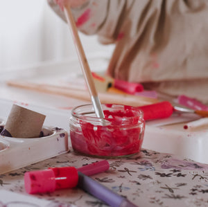 Child playing with red paint and a brush on a table with art supplies.