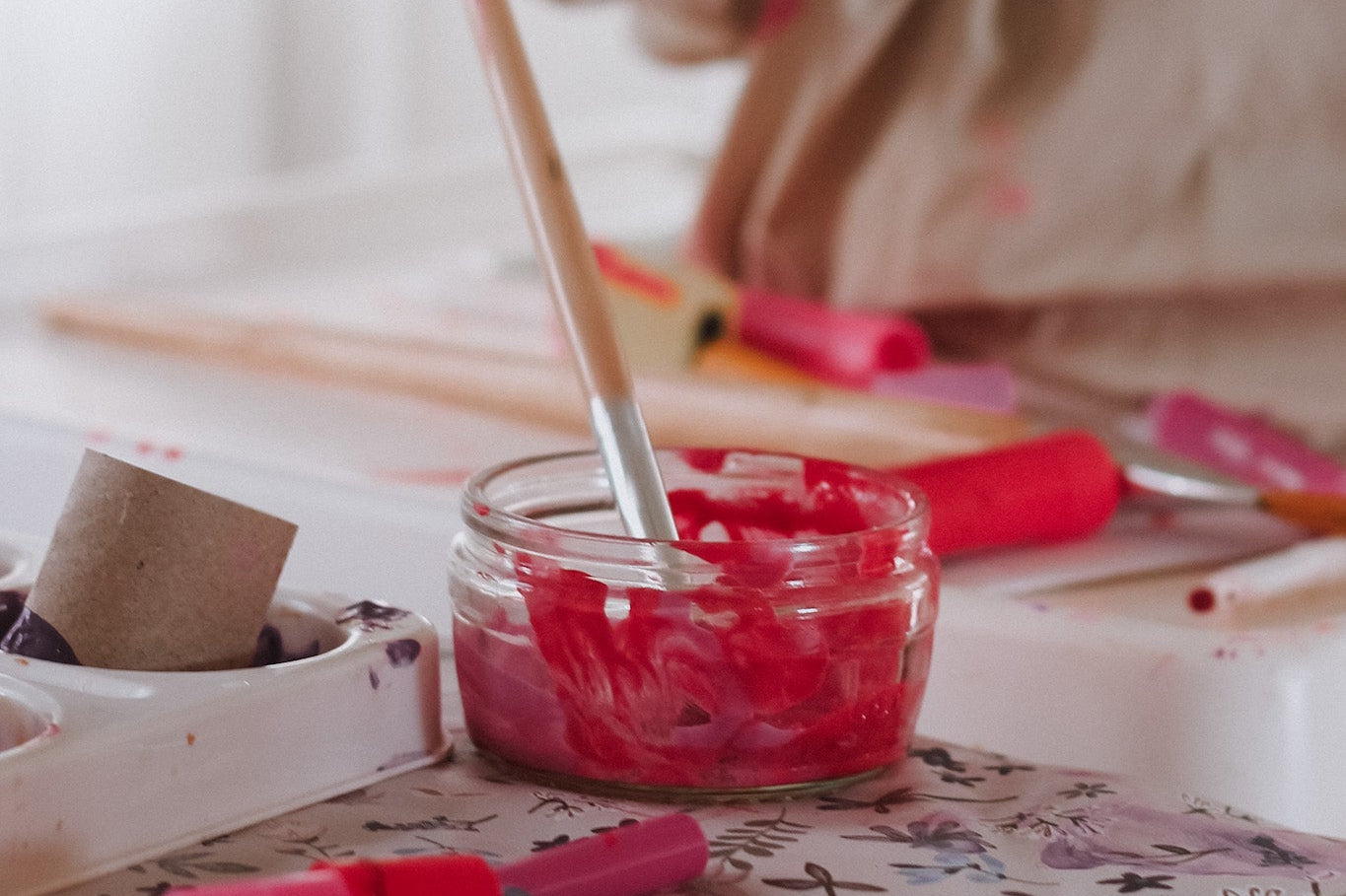 Child playing with red paint and a brush on a table with art supplies.
