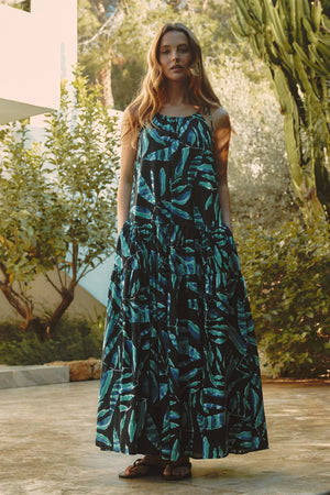 Woman in a blue and black patterned dress standing outdoors with greenery in the background