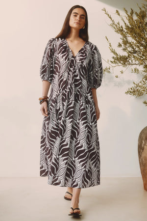 Woman wearing a patterned dress standing next to a potted plant indoors.
