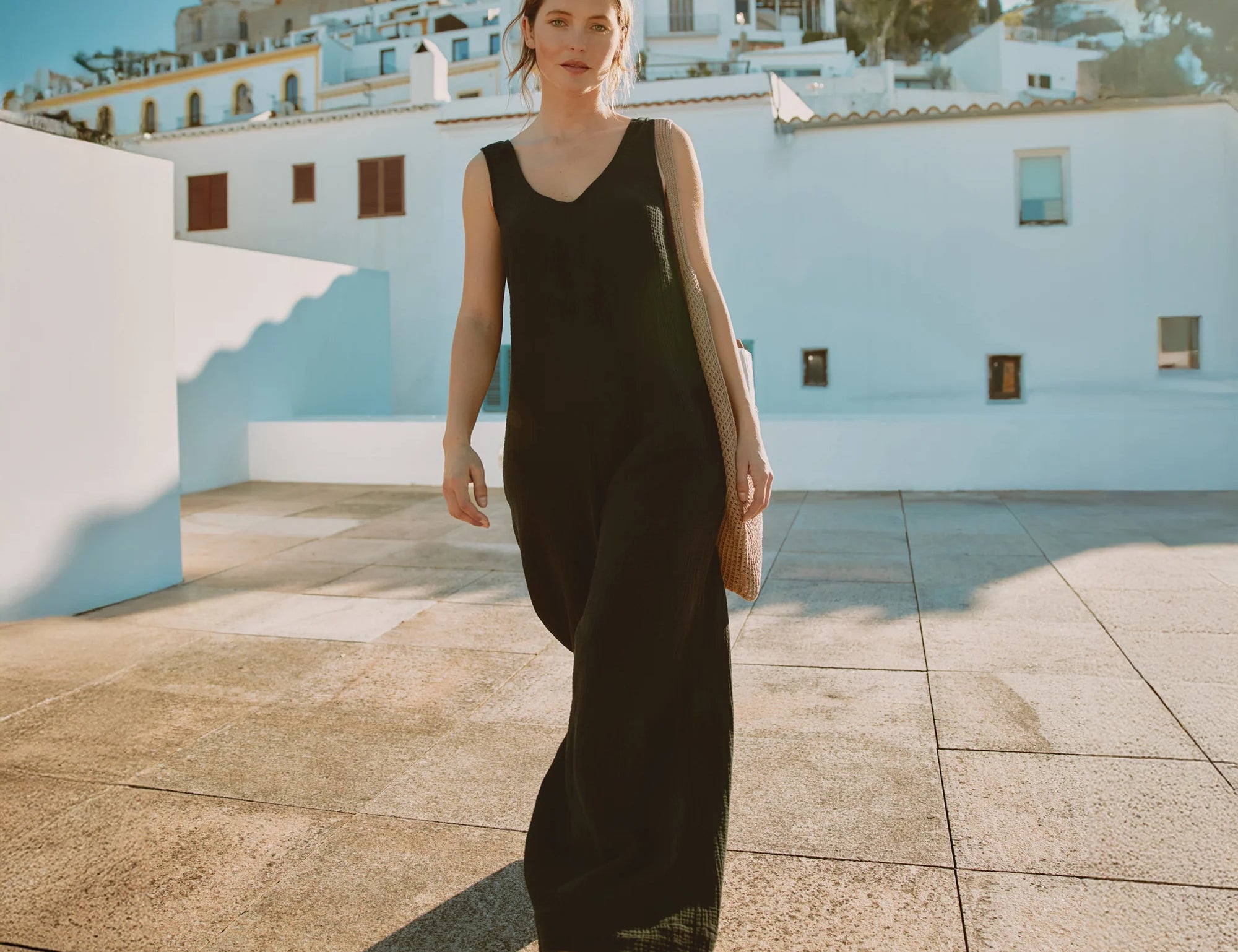 Woman in a black sleeveless jumpsuit standing on a sunlit street with white buildings in the background.