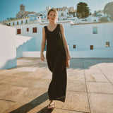 Woman in a black sleeveless jumpsuit standing on a sunlit street with white buildings in the background.