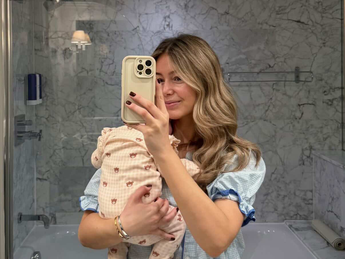 Woman holding a baby in a bathroom taking a mirror selfie.