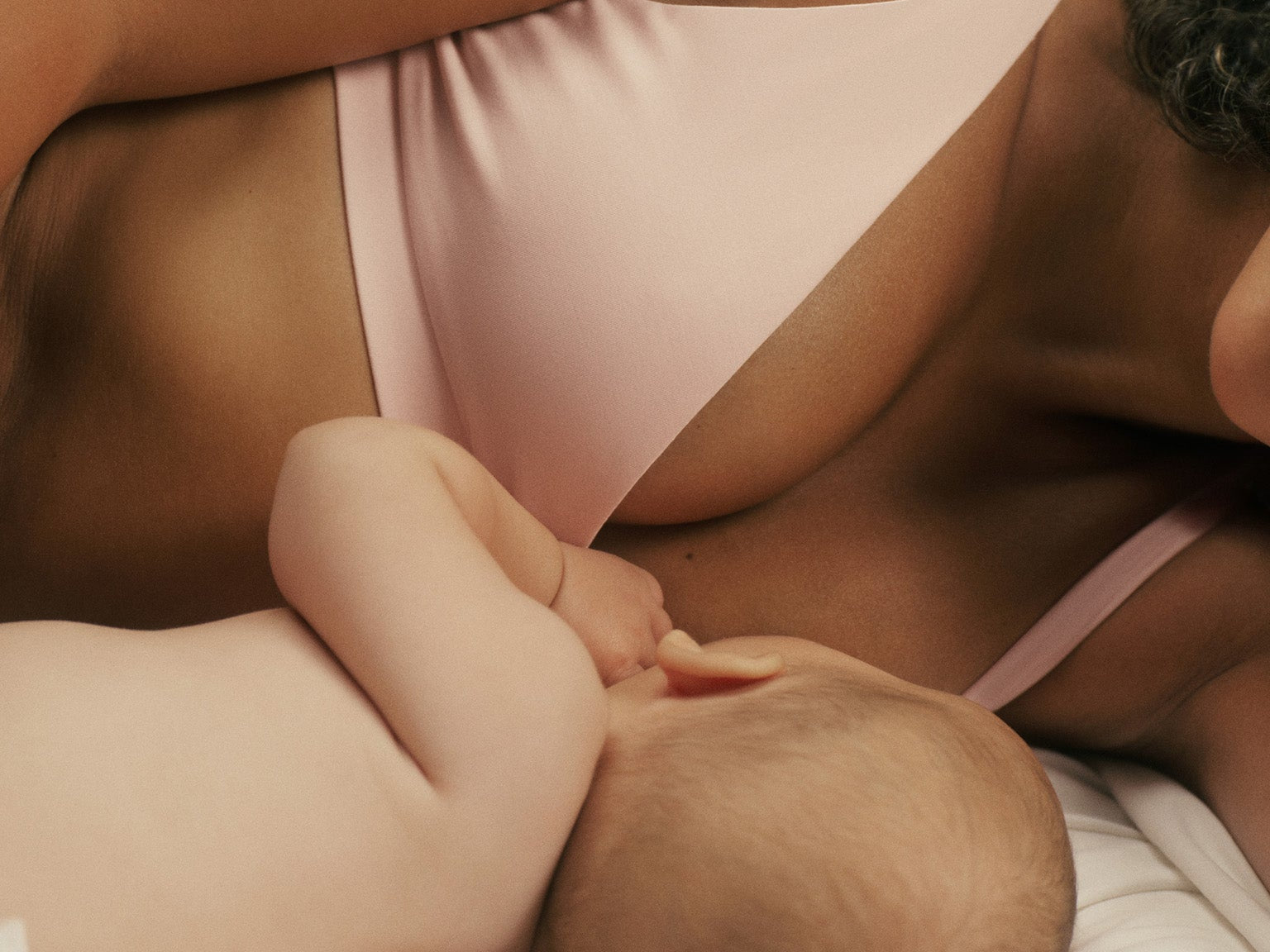 A close-up lifestyle shot of a mother breastfeeding her baby while wearing a light pink nursing bra and matching nursing bottoms on a white bed.