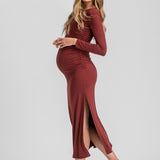 A pregnant woman modeling a long-sleeve, ruched maternity maxi dress in a deep burgundy color, paired with dark brown slingback heels, against a light gray studio background.