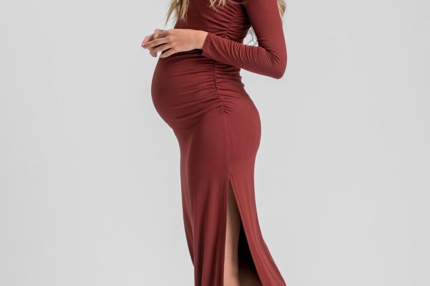 A pregnant woman modeling a long-sleeve, ruched maternity maxi dress in a deep burgundy color, paired with dark brown slingback heels, against a light gray studio background.