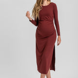 A woman modeling a long-sleeve, gathered maternity dress in a deep burgundy color, standing against a light gray studio background.
