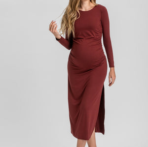 A woman modeling a long-sleeve, gathered maternity dress in a deep burgundy color, standing against a light gray studio background.
