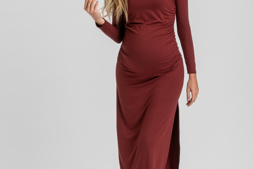 A woman modeling a long-sleeve, gathered maternity dress in a deep burgundy color, standing against a light gray studio background.