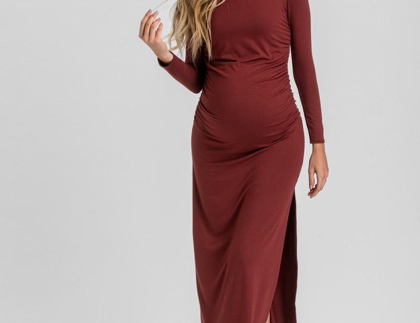 A woman modeling a long-sleeve, gathered maternity dress in a deep burgundy color, standing against a light gray studio background.