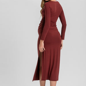 A woman modeling a long-sleeve, ruched midi dress in a deep burgundy or rust red color, shown from a three-quarters back view against a light gray studio background.