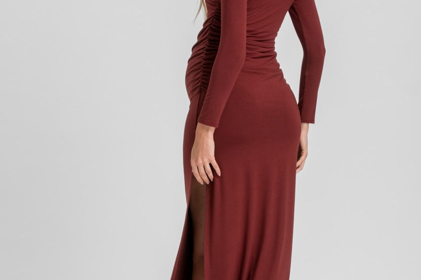 A woman modeling a long-sleeve, ruched midi dress in a deep burgundy or rust red color, shown from a three-quarters back view against a light gray studio background.