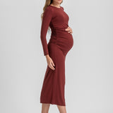 A pregnant woman modeling a long-sleeve, ruched maternity dress in a deep burgundy color, paired with black slingback kitten heels, against a light gray studio background.