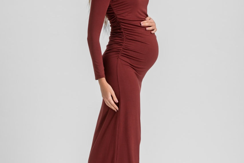 A pregnant woman modeling a long-sleeve, ruched maternity dress in a deep burgundy color, paired with black slingback kitten heels, against a light gray studio background.