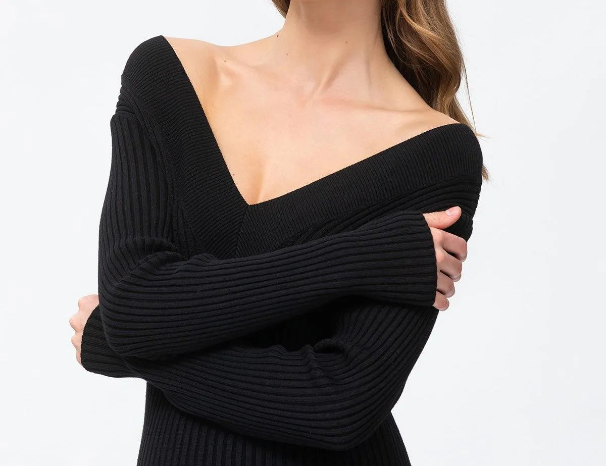 A woman modeling a black ribbed knit off-the-shoulder sweater dress against a plain white studio background.