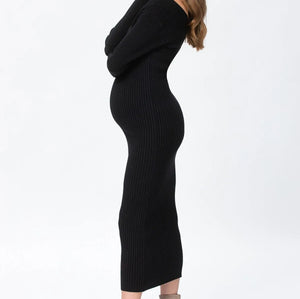 A woman in a side profile wearing a black ribbed knit long-sleeve midi dress and beige ankle boots, standing against a plain white studio background.