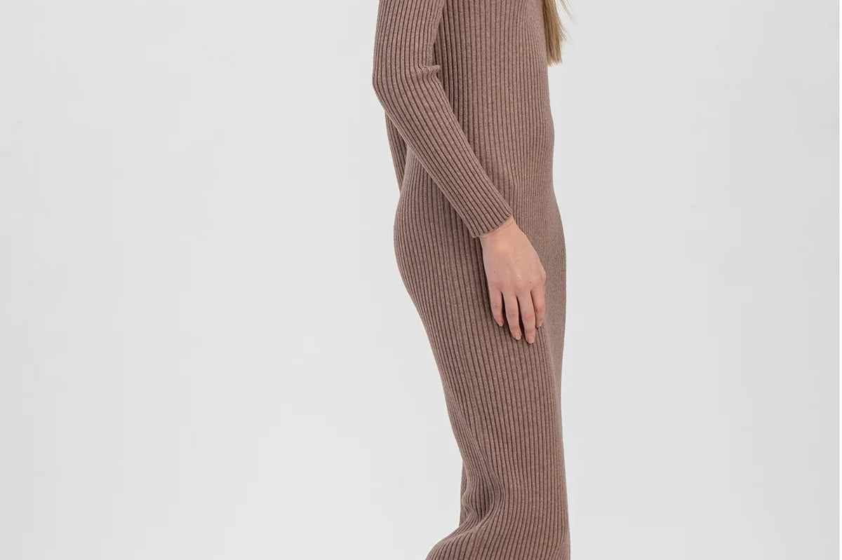 A woman modeling a ribbed knit long-sleeve midi dress in taupe brown, paired with dark brown patent leather ankle boots, against a light gray studio background.